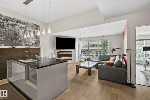 Open-concept living area featuring wood-finish flooring, a contemporary kitchen island with an integrated sink and dark countertop, a built-in fireplace, and a private balcony - 201 10388 105 Street, Edmonton, AB 