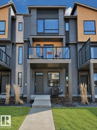 Modern townhome exterior featuring multi-toned siding, large windows, an upper-level balcony with glass railings, a concrete walkway, and manicured lawn - 311 32 Street, Edmonton, AB 