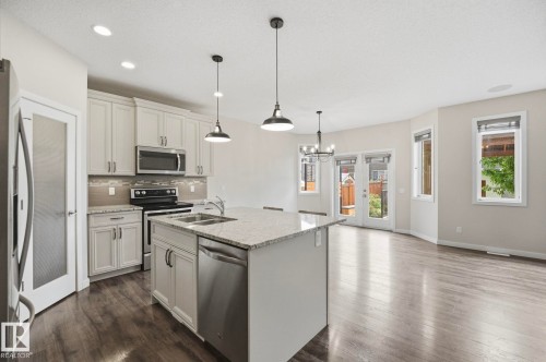 Open-concept living space featuring wood-finish flooring, a kitchen island with a double sink and pendant lighting, light-colored cabinetry, stainless steel appliances, and a tiled backsplash - 33 Riviere Terrace, St. Albert, AB 