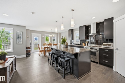 Modern kitchen featuring dark cabinetry, stainless steel appliances, a large center island with seating, pendant lighting, and wood-finish flooring - 5430 Edworthy Way, Edmonton, AB - Indoor Photo Showing Other Room