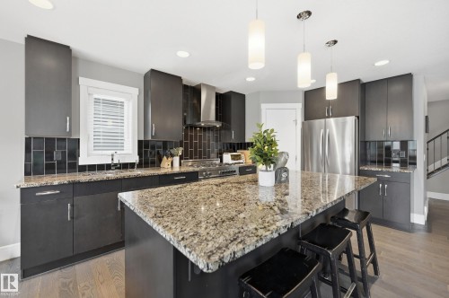 Modern kitchen featuring a large granite-topped island with pendant lighting, stainless steel appliances, dark cabinetry, and black subway tile backsplash - 5430 Edworthy Way, Edmonton, AB - Indoor Photo Showing Kitchen With Stainless Steel Kitchen With Upgraded Kitchen