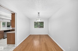 Open-concept living space featuring wood-finish flooring, a contemporary ceiling light fixture, a window with exterior greenery, and a partial view of a kitchen with wood cabinetry and tile flooring - 