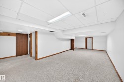 Spacious lower-level room featuring light-toned carpeting, white walls, wood-finish trim, and a drop ceiling with integrated fluorescent lighting - 