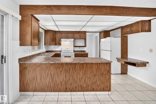 Spacious kitchen featuring extensive wood cabinetry, white tile flooring, a U-shaped countertop configuration, a built-in breakfast bar, and white appliances - 9843 188 Street, Edmonton, AB 