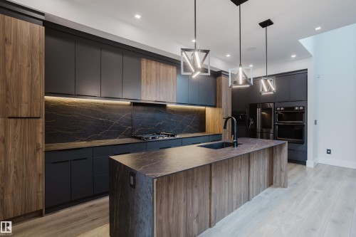 Modern kitchen featuring a waterfall island with an undermount sink and matte black faucet, dark flat-panel cabinetry, wood-finish accent cabinetry, and a black slab backsplash with veining - 1313 Clement Crest, Edmonton, AB 