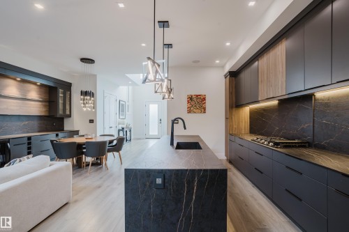 Open-concept kitchen featuring an oversized island with an undermount sink, dark cabinetry with integrated appliances, and light wood-finish flooring throughout - 1313 Clement Crest, Edmonton, AB 