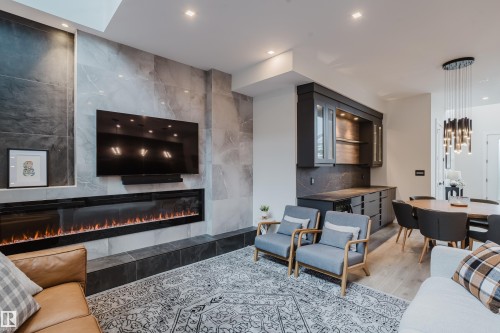 Living area featuring a floor-to-ceiling tiled fireplace surround, an elongated electric fireplace, a wall-mounted television, built-in cabinetry with glass doors, and recessed lighting - 1313 Clement Crest, Edmonton, AB 