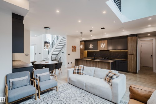 Open-concept living space featuring light wood-finish flooring, recessed lighting, a modern kitchen with dark cabinetry and a patterned backsplash, an island with pendant lighting, and a dining area with a contemporary chandelier - 1313 Clement Crest, Edmonton, AB 
