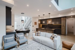 Open-concept living space featuring light wood-finish flooring, recessed lighting, a modern kitchen with dark cabinetry and a patterned backsplash, an island with pendant lighting, and a dining area with a contemporary chandelier - 