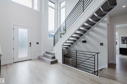 Grand entryway featuring a floating staircase with sleek horizontal iron railings, high ceilings, large windows, wood-finish flooring, and a frosted glass panel entry door - 1313 Clement Crest, Edmonton, AB 