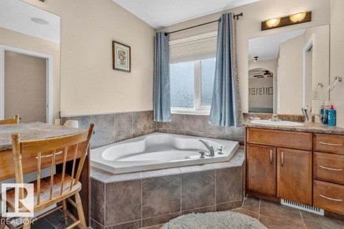Corner soaking tub with tiled surround, single vanity with integrated sink, wood-finish cabinetry, and framed mirror with overhead lighting - 39 Wilkinson Place, Leduc, AB 