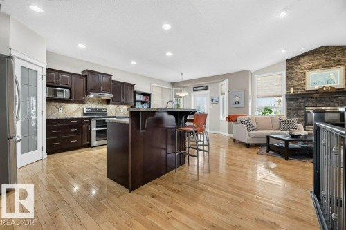 Open-concept living area featuring a kitchen with dark wood cabinetry and stainless steel appliances, a center island with seating, natural wood-finish flooring, and a stone-faced fireplace - 5528 43 Street, Lamont, AB 