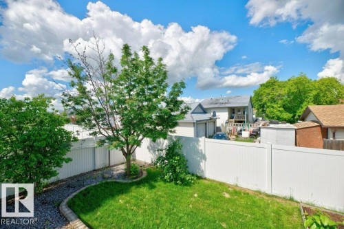 Enclosed backyard with a white privacy fence, featuring a manicured lawn, established trees, and a gravel-filled garden bed with a curved stone border - 5528 43 Street, Lamont, AB 