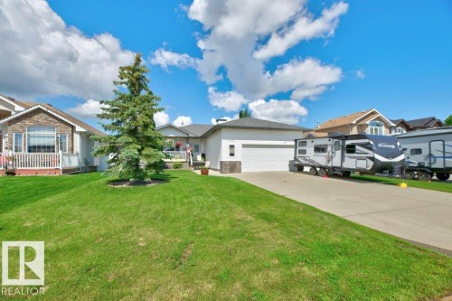 Front exterior featuring a stone and stucco facade, an attached garage, a mature evergreen tree, a concrete driveway, and a manicured lawn - 5528 43 Street, Lamont, AB 