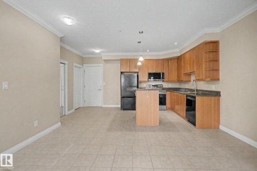 Open-concept kitchen and living area featuring warm wood cabinetry, dark countertops, black appliances, a central island, and tile flooring throughout - 413 10116 80 Avenue, Edmonton, AB 