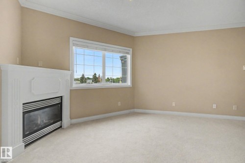 Neutral-toned room featuring a gas fireplace with a white mantel, large grid-patterned window, crown molding, and light-colored carpeting - 413 10116 80 Avenue, Edmonton, AB 