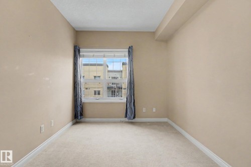Bright room featuring neutral carpet flooring, light beige walls, white baseboards, and a prominent window - 413 10116 80 Avenue, Edmonton, AB 