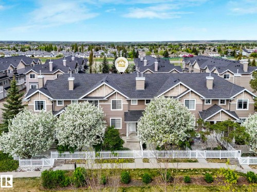Townhome exterior featuring tan siding, dark shingle roofing, white trim, white picket fencing, and mature flowering trees - 62 3075 Trelle Crescent Nw, Edmonton, AB - Outdoor With Facade