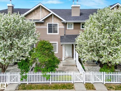 Townhome exterior featuring tan siding, decorative shake accents, a dark shingle roof, a white picket fence, and mature blooming trees - 62 3075 Trelle Crescent Nw, Edmonton, AB - Outdoor