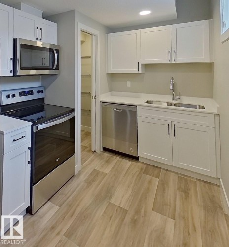 Kitchen featuring white shaker-style cabinetry, stainless steel appliances, a double basin sink, light-toned countertops, and wood-finish flooring - #Basement 10923 159 St Nw, Edmonton, AB - Indoor Photo Showing Kitchen With Stainless Steel Kitchen With Double Sink