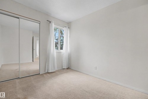 Room featuring neutral-toned carpeting, a window with white curtains, white walls, and a mirrored closet with sliding doors - 198 11421 34 Street, Edmonton, AB 