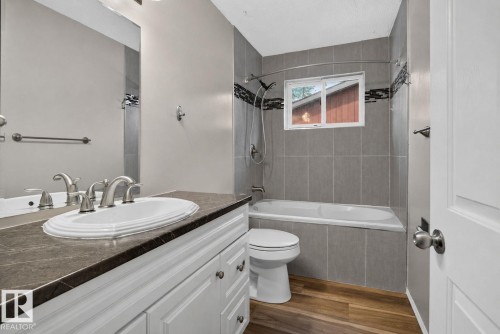 Bathroom featuring a white vanity with a dark countertop and undermount sink, brushed nickel fixtures, a tub-shower combination with gray tile surround, and wood-finish flooring - 13431 109 Street, Edmonton, AB 
