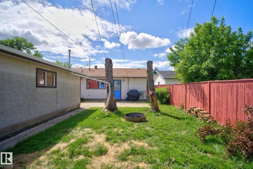 Expansive backyard featuring a red wood fence, established trees, a grass lawn, and a round fire pit with stone surround - 13431 109 Street, Edmonton, AB 