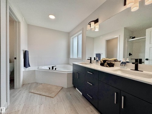 Bathroom featuring an integrated corner bathtub with white tile surround, a dark wood-finish double vanity with white countertops and matte black fixtures, light wood-finish flooring, and a large wall-mounted mirror - 33 Sumac Close, Fort Saskatchewan, AB 