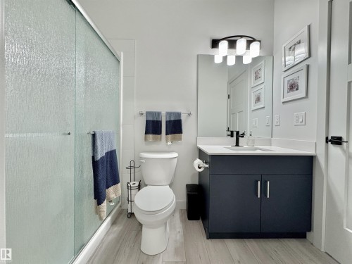 Bathroom featuring a textured glass shower enclosure, a dark vanity with a white countertop, wood-finish flooring, a contemporary mirror, and a modern wall-mounted light fixture - 33 Sumac Close, Fort Saskatchewan, AB 