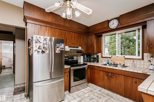 Kitchen featuring wood-paneled cabinetry, stainless steel appliances, a double basin sink, and a tiled countertop with coordinating backsplash - 14231 80 Street, Edmonton, AB 