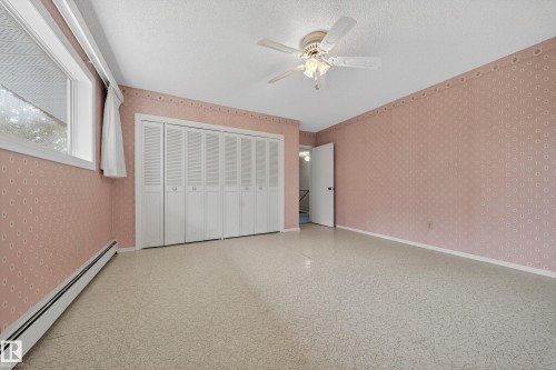 Spacious room with patterned wallpaper, ceiling fan, wide window, baseboard heating, and ample closet space with bi-fold louvered doors - 707 65 Avenue, Edmonton, AB 