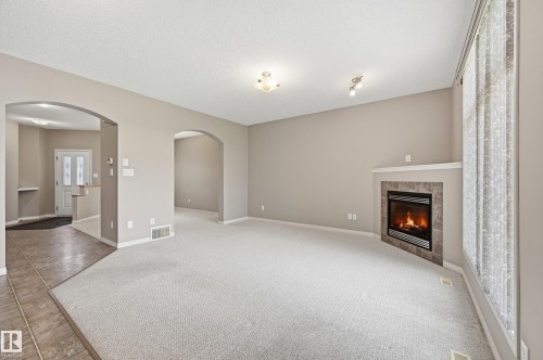 Spacious room featuring a fireplace with a tiled surround and mantel, light-toned carpet, and a large window with sheer curtains - 7460 Singer Landing, Edmonton, AB 