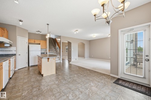 Open-concept kitchen and living area featuring tile flooring, wooden cabinetry, a kitchen island with countertop, and an exterior door with integrated blinds - 7460 Singer Landing, Edmonton, AB 