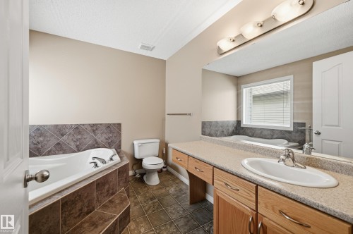 Bathroom featuring a built-in jetted tub with tile surround, a single vanity with an integrated sink and light wood cabinetry, and a mirrored wall with strip lighting - 7460 Singer Landing, Edmonton, AB 