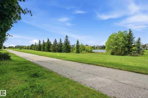 Paved pathway bordered by manicured green space, mature evergreen trees, and a distant body of water - 7460 Singer Landing, Edmonton, AB 