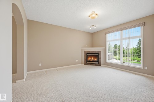 Corner fireplace with a stone surround and mantel, large window with vertical blinds, textured carpeting, neutral wall tones, and arched entryway - 7460 Singer Landing, Edmonton, AB 