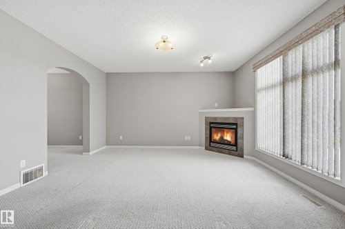 Carpeted room featuring a corner fireplace with a tiled surround, a large window with vertical blinds, an arched doorway, and light gray wall paint - 7460 Singer Landing, Edmonton, AB 