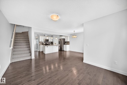 Open-concept living area featuring dark wood-finish flooring, a kitchen with white cabinetry and stainless steel appliances, a kitchen island with seating, a carpeted staircase, and an additional dining area with a decorative chandelier - 2433 Glenridding Boulevard, Edmonton, AB 