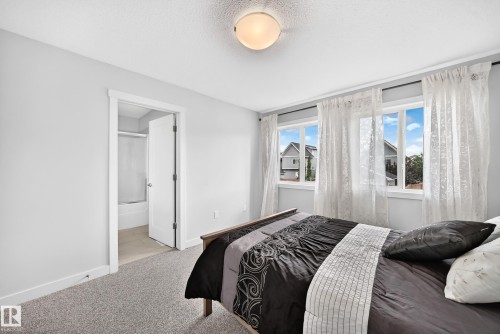 Carpeted bedroom with light gray walls, featuring two large windows with white trim, a flush-mount ceiling light, and an open doorway leading to a bathroom with a shower-over-tub combination - 2433 Glenridding Boulevard, Edmonton, AB 