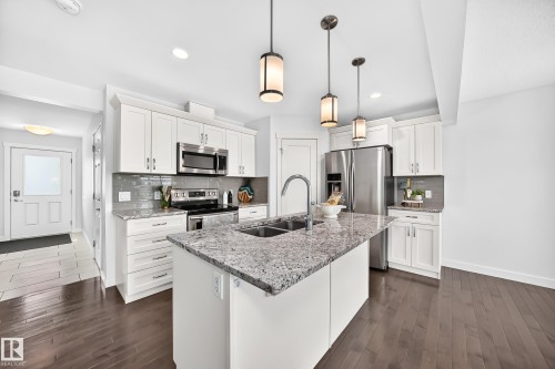 Kitchen featuring a central island with a double basin sink, stainless steel appliances, white cabinetry, a subway tile backsplash, and dark wood-finish flooring - 2433 Glenridding Boulevard, Edmonton, AB 