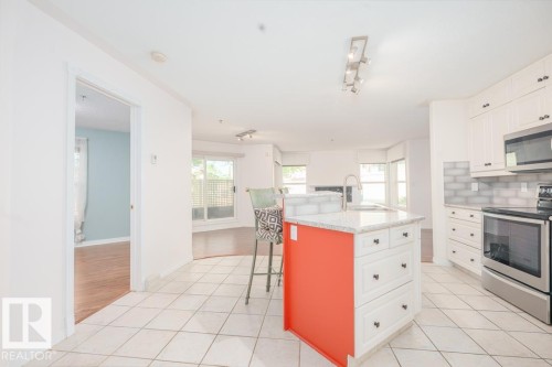 Spacious kitchen featuring white cabinetry, light-toned tiled flooring, stainless steel appliances, a center island with a granite-style countertop, and a subway tile backsplash - 108 12028 103 Avenue, Edmonton, AB 