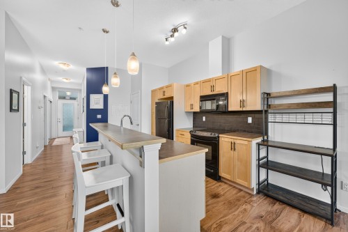 Kitchen with wood-finish flooring, natural wood cabinetry, a breakfast bar with pendant lighting, a dark tile backsplash, and black appliances - 410 10531 117 Street, Edmonton, AB 