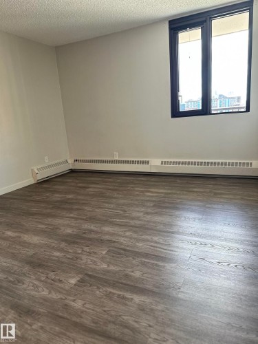 Spacious room with wood-finish flooring, a large window with black trim, and light neutral wall paint - 706 10145 109 Street, Edmonton, AB 