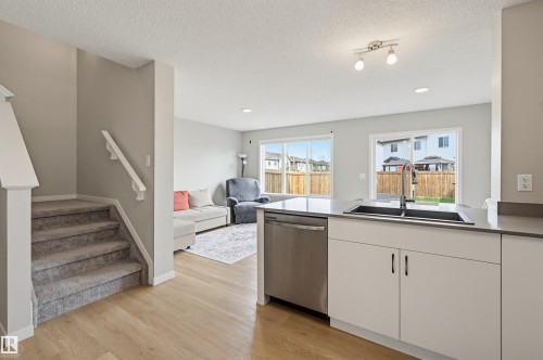 Open-concept kitchen and living area featuring light wood-finish flooring, a kitchen island with a double basin sink and stainless steel dishwasher, white cabinetry, and a carpeted staircase with white handrails - 56 Springbrook Wynd, Spruce Grove, AB - Indoor Photo Showing Kitchen With Double Sink