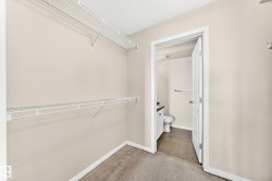 Walk-in closet featuring wire shelving, light neutral walls, and a carpeted floor, with an entryway to a bathroom featuring a toilet, vanity, and tiled flooring - 