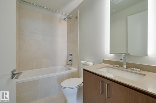 Modern bathroom featuring a wood-finish vanity with a light-toned countertop and integrated sink, an illuminated mirror, and a full-size bathtub with polished chrome fixtures and light-toned tile surround - #3901 10360 102 St Nw Nw, Edmonton, AB - Indoor Photo Showing Bathroom