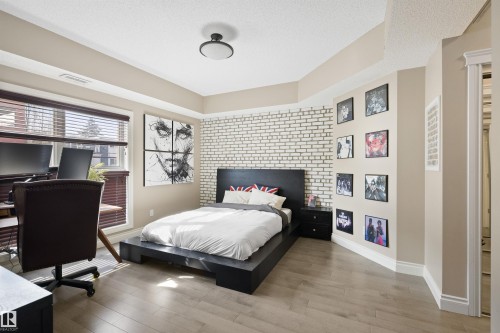 Spacious room featuring light wood-finish flooring, a prominent white brick accent wall, and a large window with horizontal blinds - #206 10232 115 St Nw, Edmonton, AB 