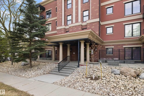 Building exterior featuring a brick facade with light-colored accent banding, a covered entryway with classical columns, and a concrete staircase with black handrails - #206 10232 115 St Nw, Edmonton, AB 