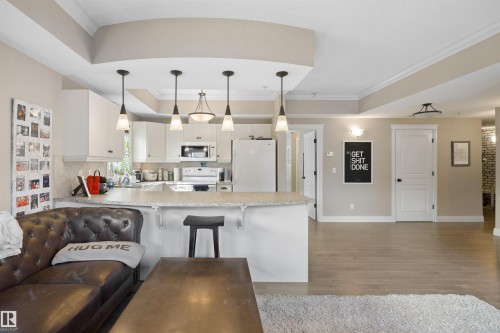 Open-concept living space featuring a kitchen with white cabinetry and a breakfast bar, recessed ceiling details, light wood-finish flooring, and white paneled doors - #206 10232 115 St Nw, Edmonton, AB 