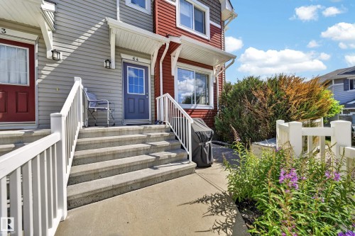 Townhouse exterior featuring a concrete walkway, light gray siding, and a contrasting red upper facade - 64 655 Tamarack Road, Edmonton, AB 